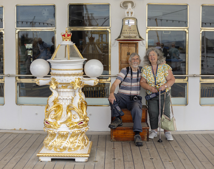 On The Royal Yacht Britannia in Edinburgh a man and woman sit under a Bell. The gold and white decorative binnacle is in the foreground.